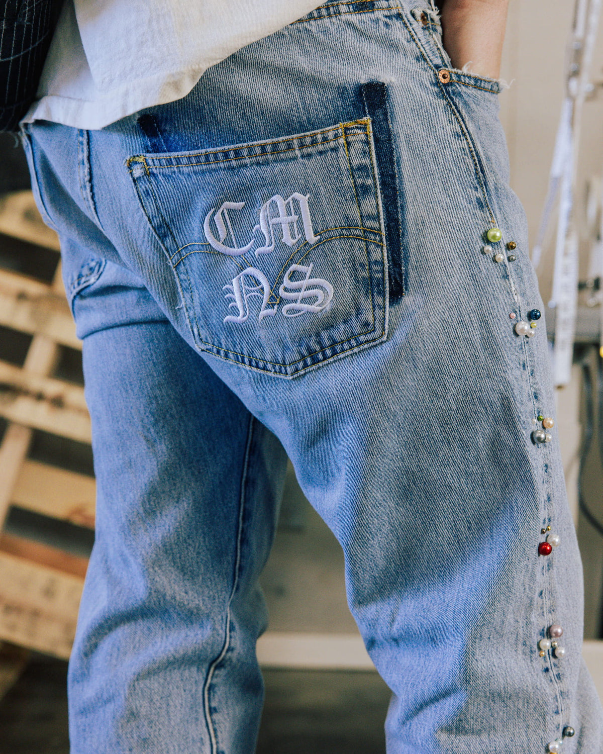 Light blue denim jeans with white embroidered letters on a back pocket and colorful bead clusters along the side seam.