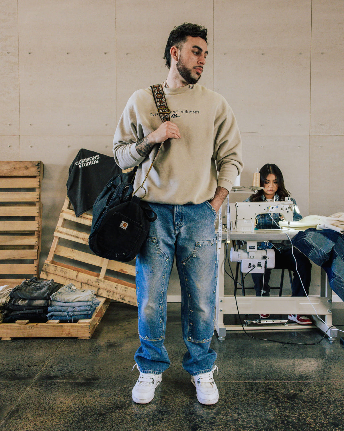 Man wearing a beige sweatshirt, blue denim carpenter jeans and white sneakers, carrying a black bag in a sewing workshop.