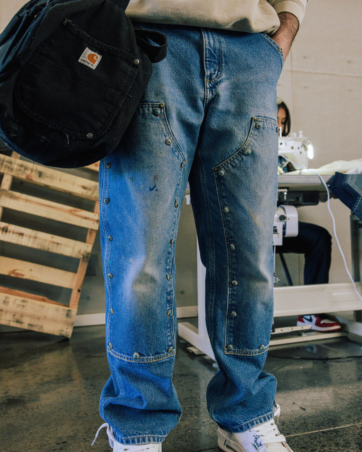 Person wearing faded blue denim jeans with studded front panels, holding a black bag in a sewing workshop.