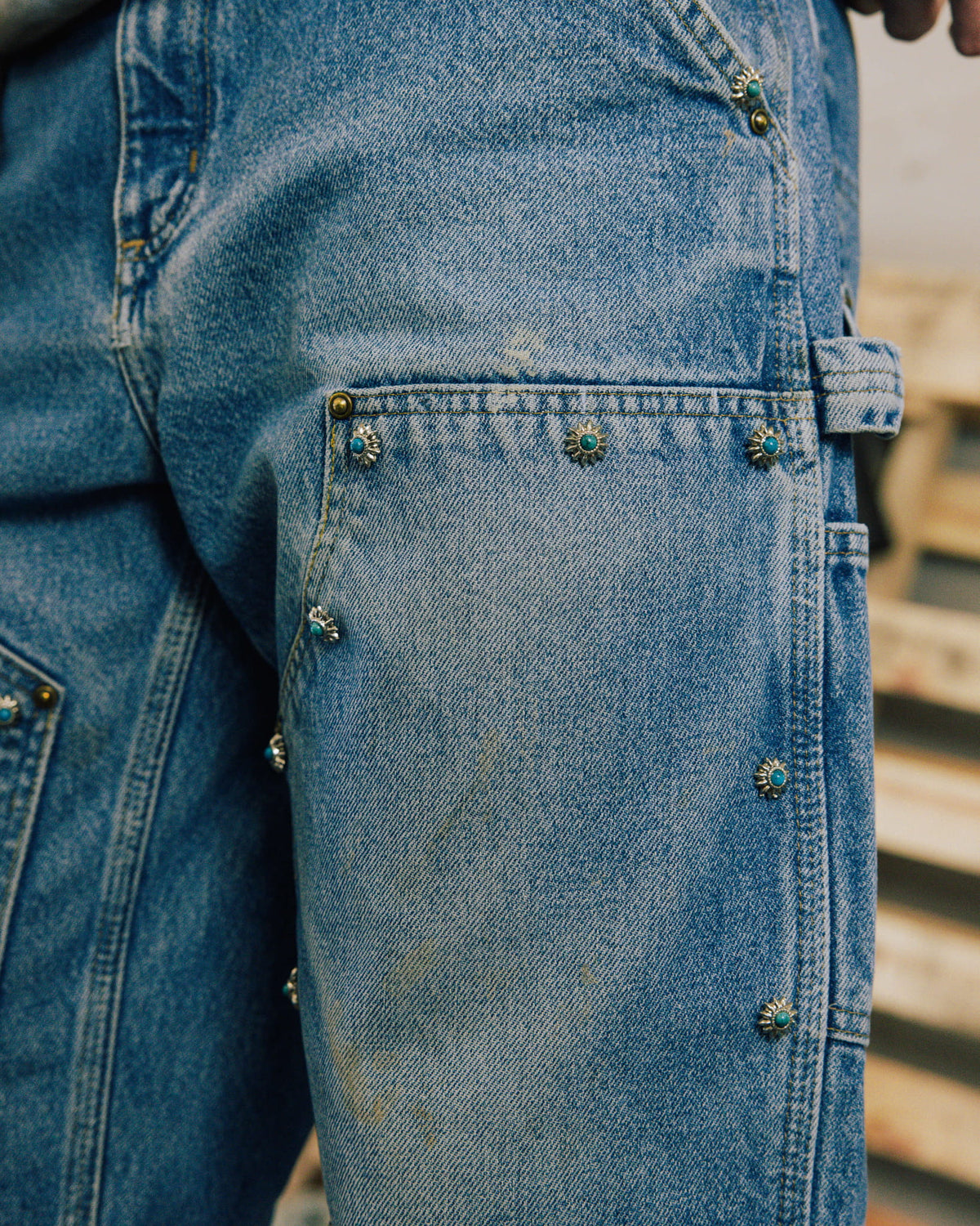 Close-up of faded blue denim carpenter jeans with turquoise-centered metal studs, reinforced panels, and a hammer loop.