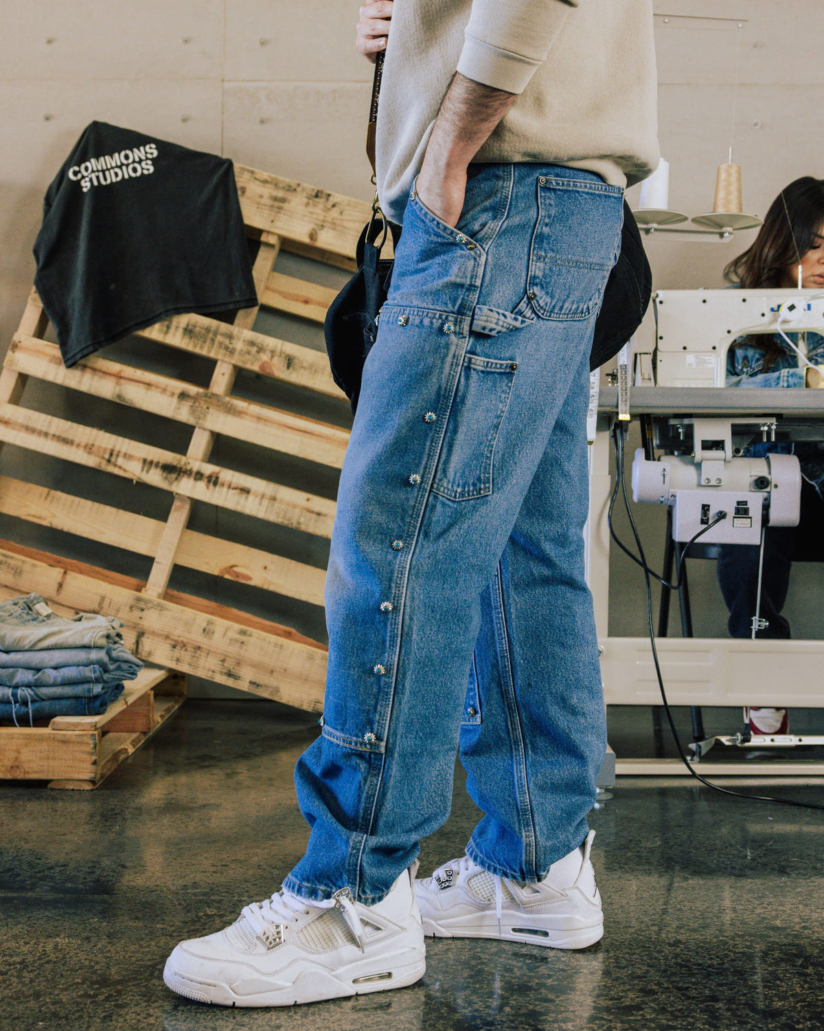Person wearing blue denim carpenter jeans with silver floral studs and white sneakers in a sewing workshop.