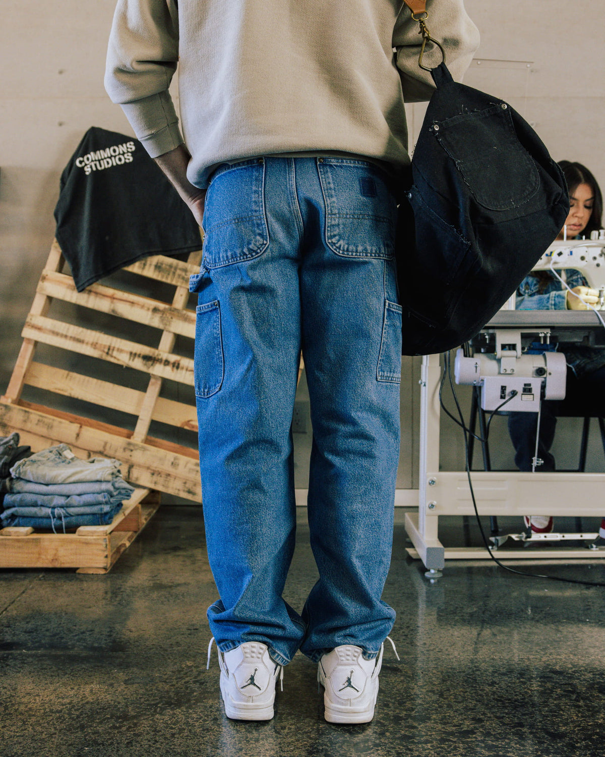 Back view of a person wearing blue denim carpenter jeans and a gray sweatshirt, carrying a black bag in a sewing workshop.