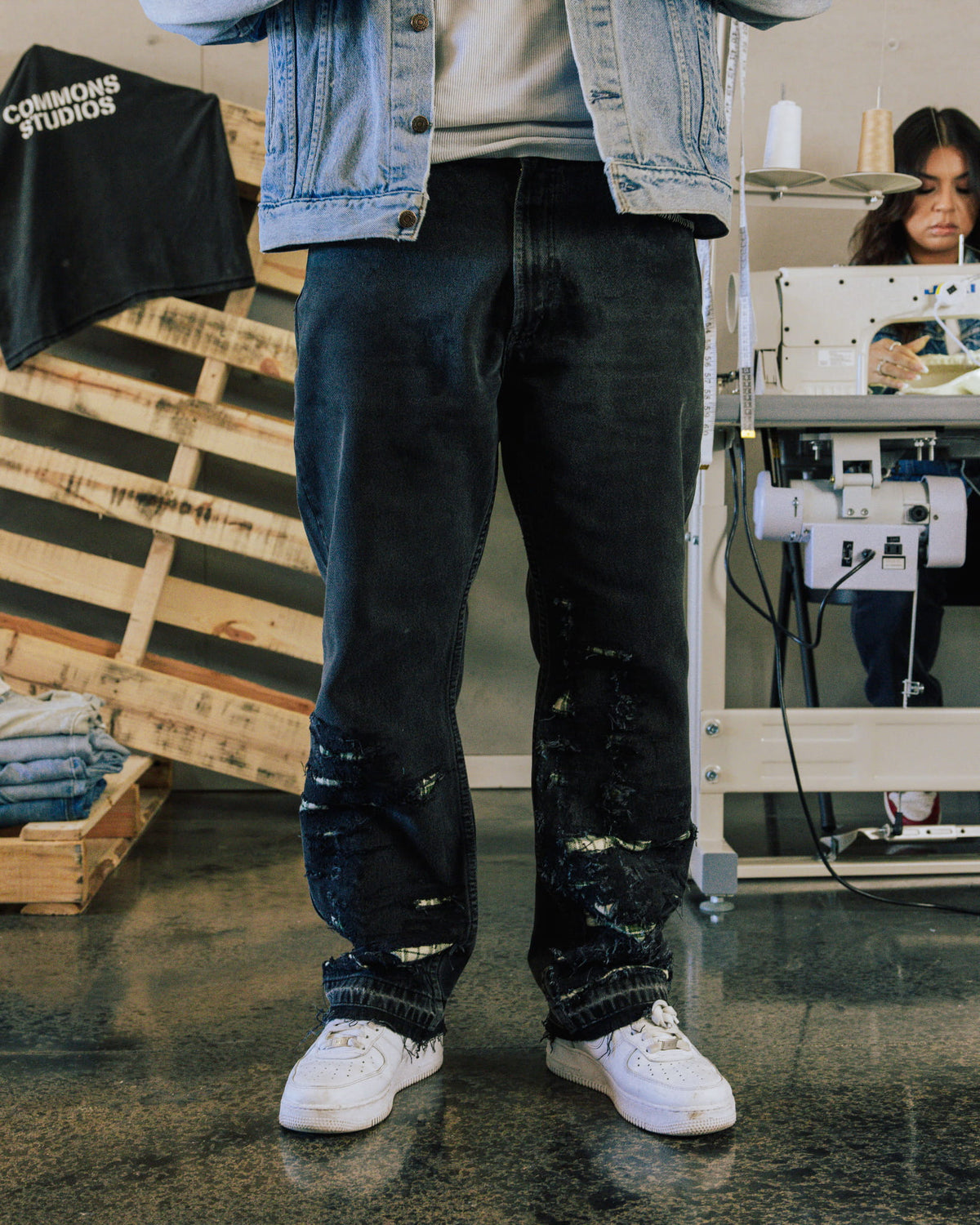Person wearing distressed dark denim jeans, a light blue denim jacket and white sneakers in a sewing workshop.