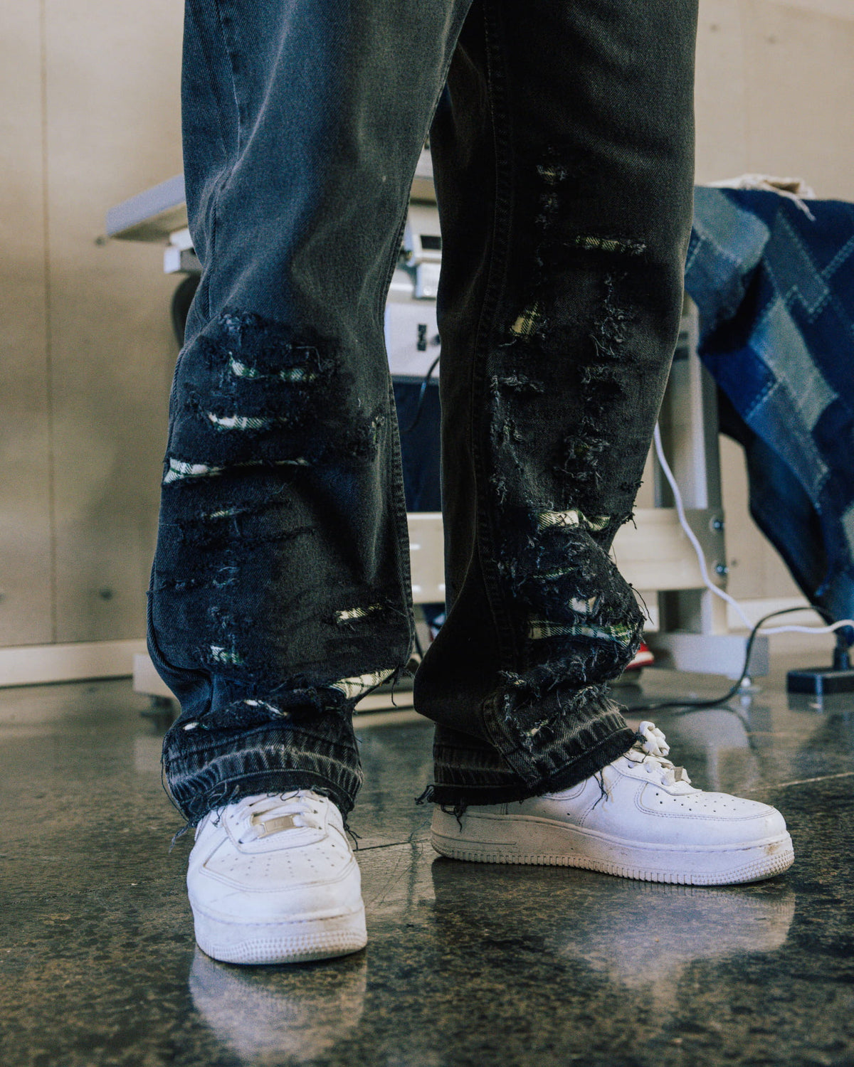 Person wearing dark blue denim jeans with heavy distressing and plaid-backed tears, paired with white sneakers indoors.