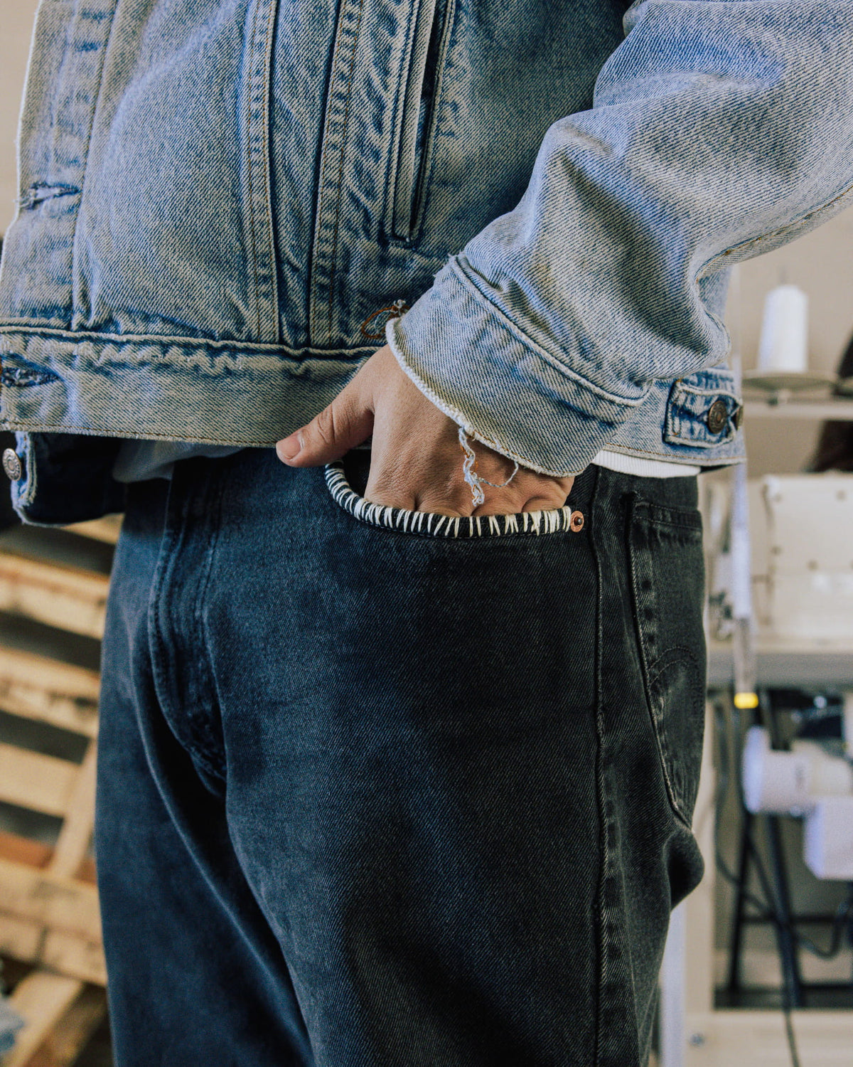 Person wearing a faded blue denim jacket and dark jeans with white stitching along the pocket, hand tucked inside.