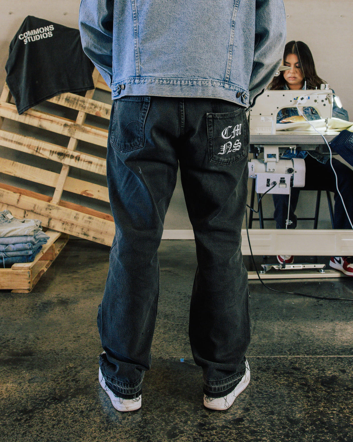 Back view of black jeans with white back-pocket lettering and a light blue denim jacket in a sewing workshop.