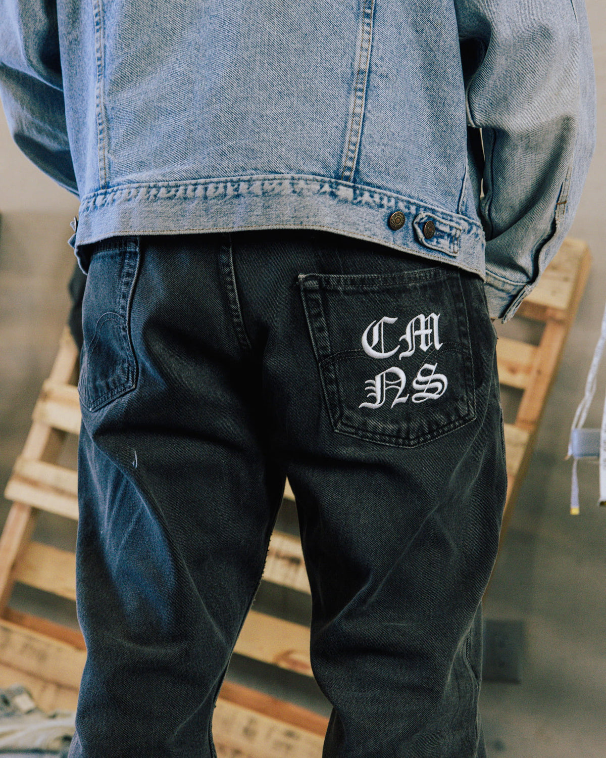 Back view of a person wearing a light blue denim jacket and dark jeans with white embroidered letters on a back pocket.
