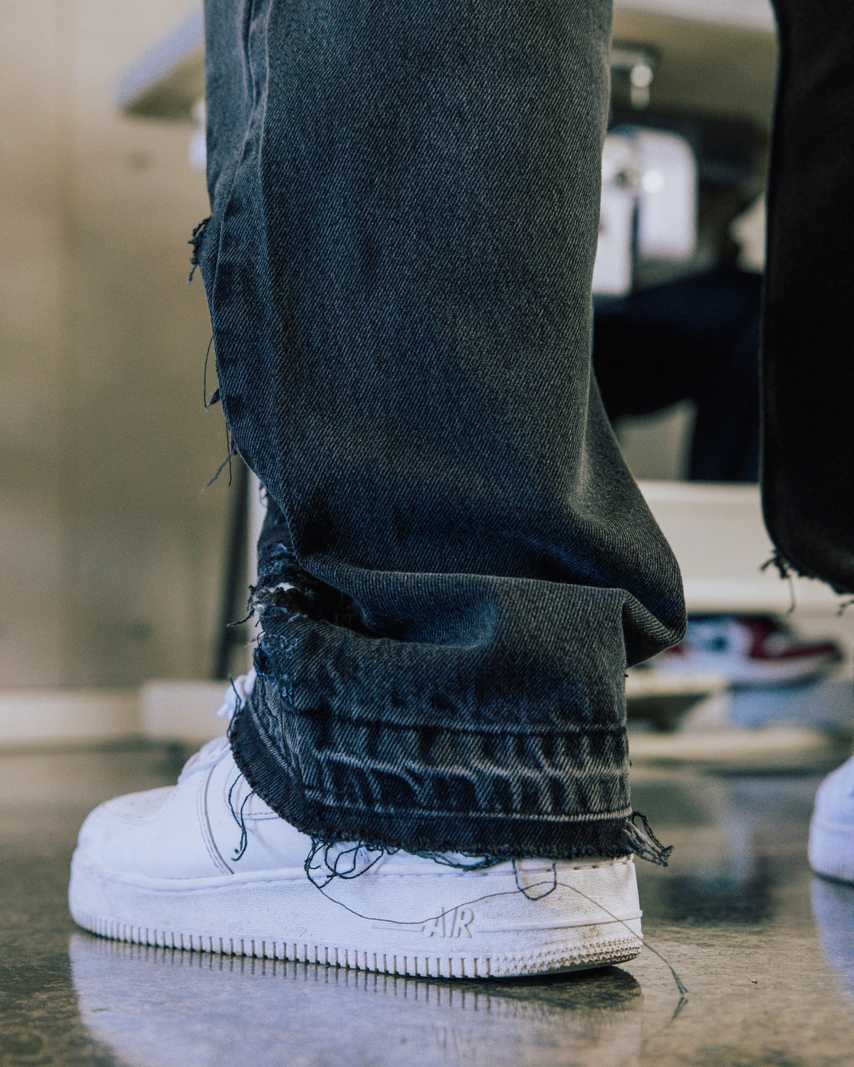 Dark blue denim jeans with frayed, distressed hems draped over white leather sneakers on a polished floor.