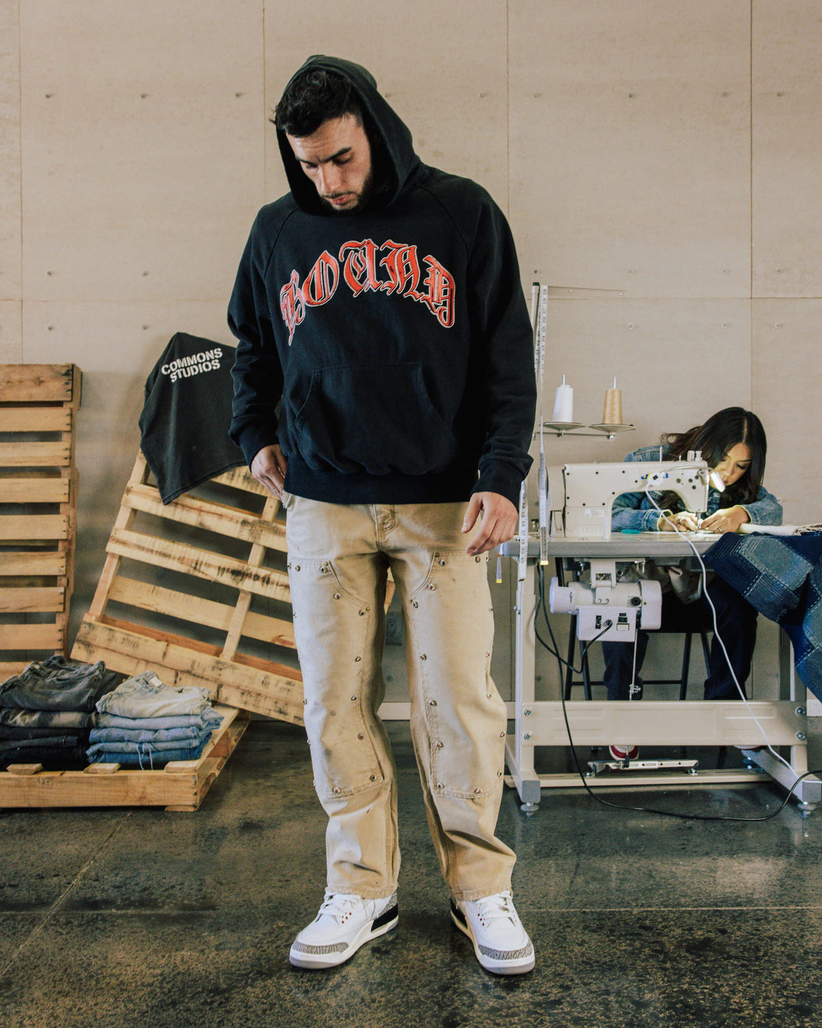 Man wearing a black hoodie with red Gothic lettering, beige studded jeans and white sneakers in a sewing workshop.