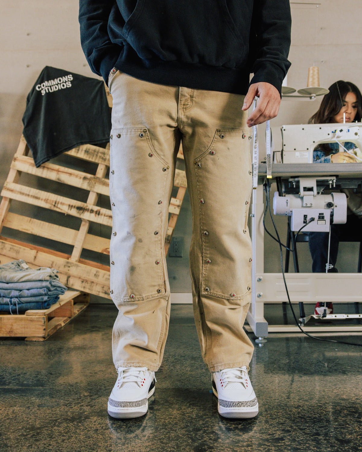Model wears beige embellished double-knee pants, a black hoodie and white sneakers in a sewing workshop.