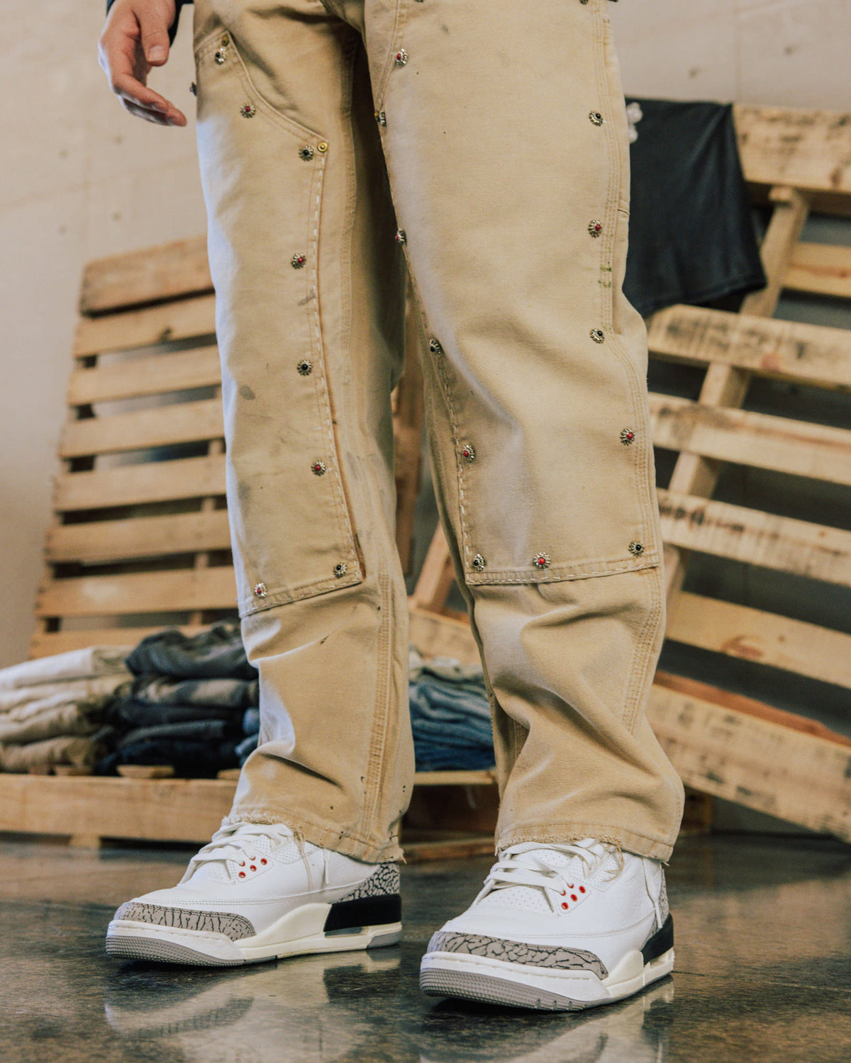 Person wearing beige pants with small jeweled studs and white sneakers, standing beside wooden pallets and folded jeans.