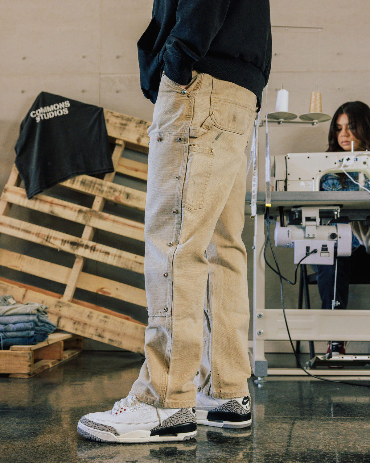 Model wears beige carpenter pants with small jeweled accents, a black sweatshirt and white sneakers in a sewing studio.
