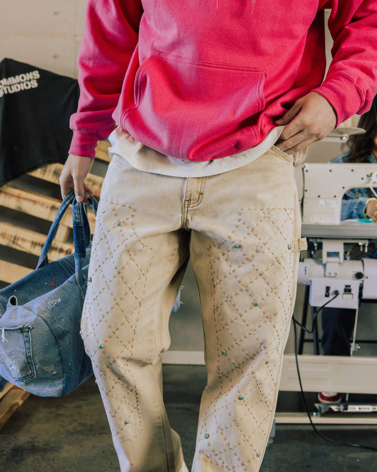 Person wearing a red hoodie and cream jeans with diamond stitching and turquoise studs, holding a blue denim bag.