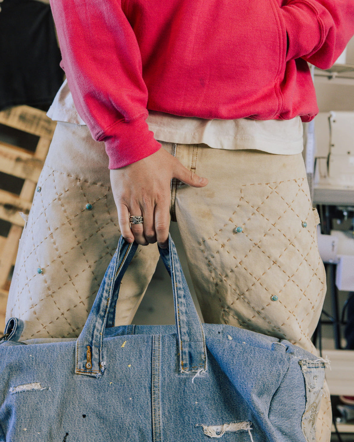Person in a red sweatshirt and beige stitched pants holding a distressed blue denim tote bag.
