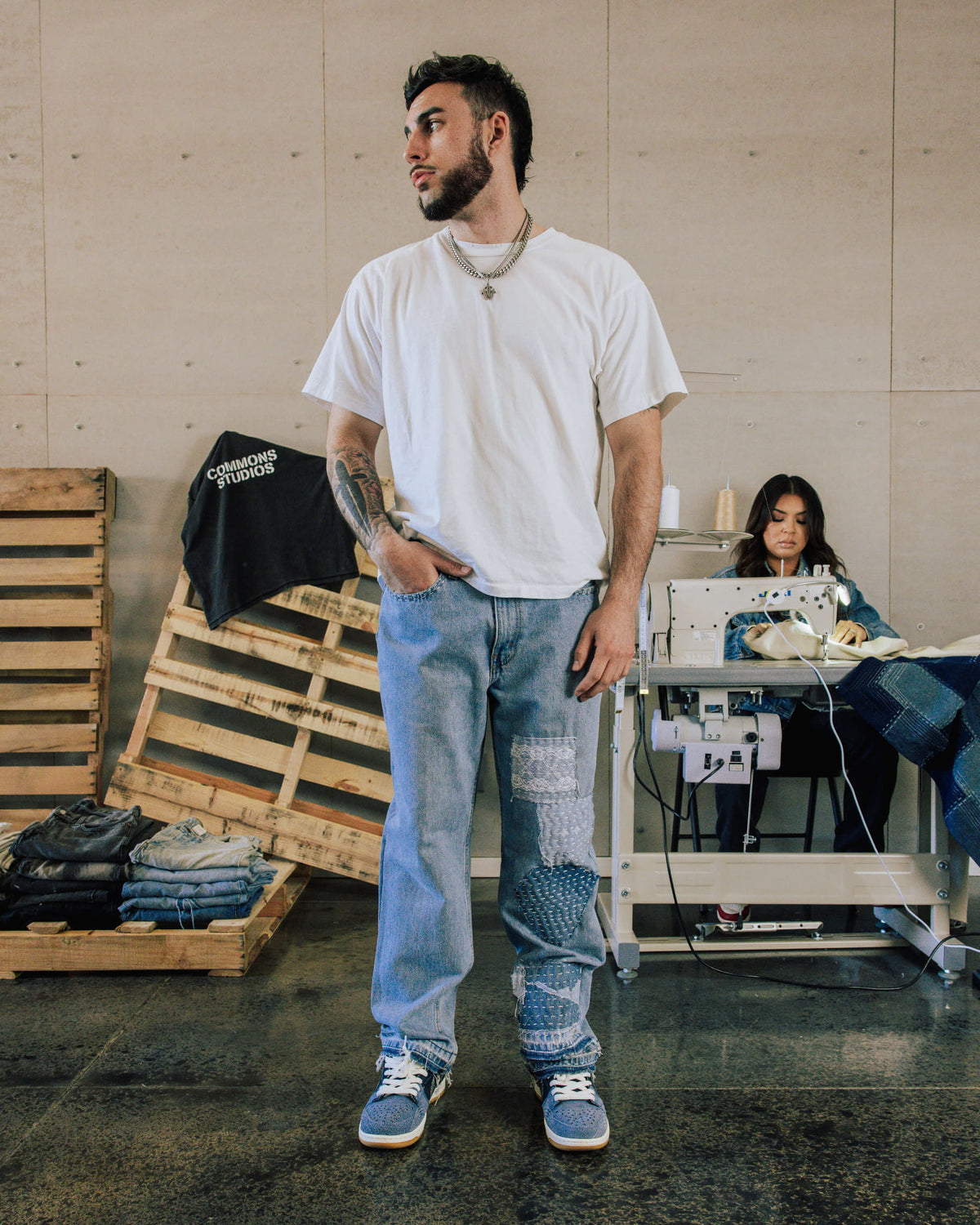 Man wearing a white T-shirt, light blue patchwork jeans and blue sneakers in a sewing workshop.