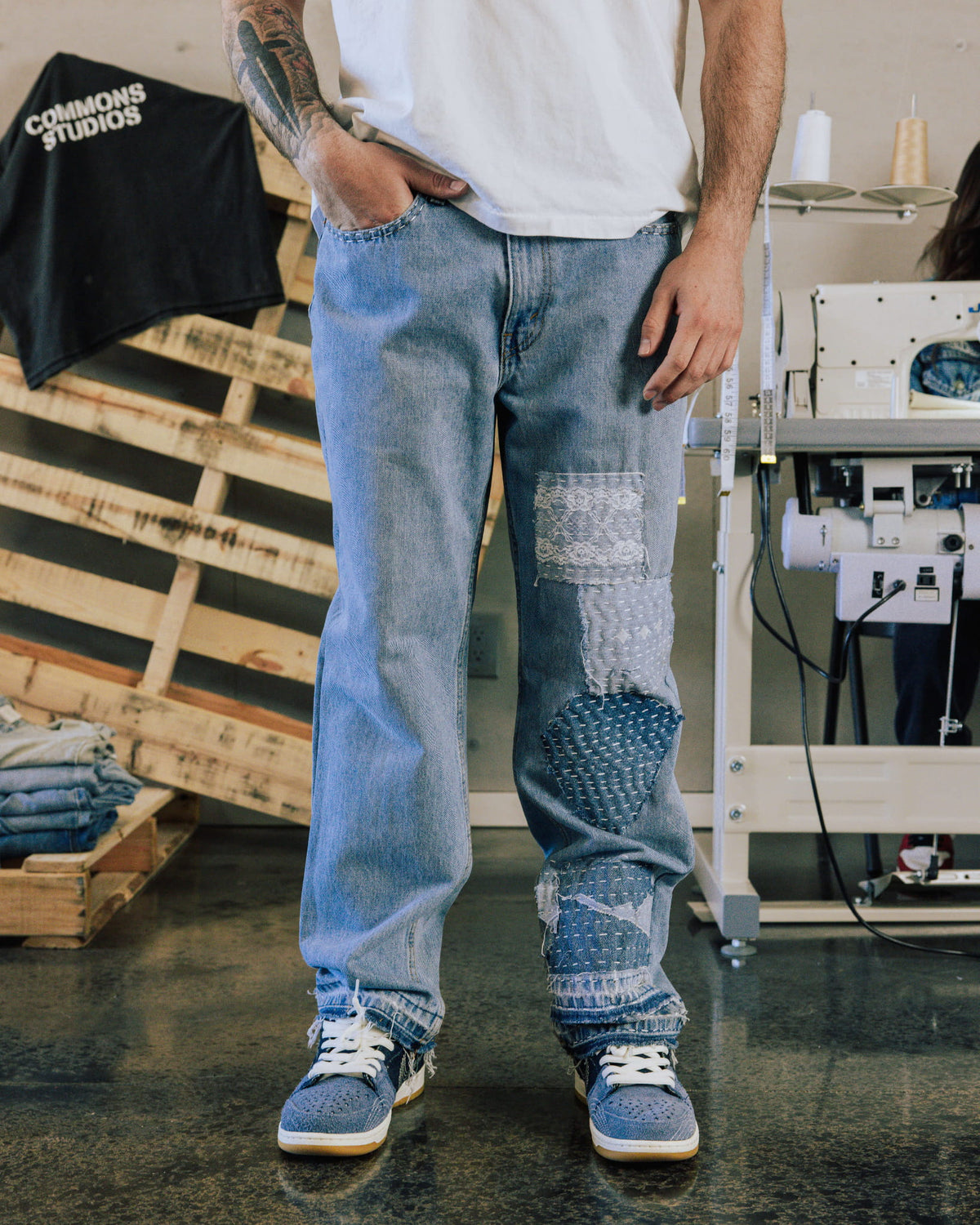 Person wearing light blue denim jeans with stitched patches, a white T-shirt and blue sneakers in a sewing workshop.