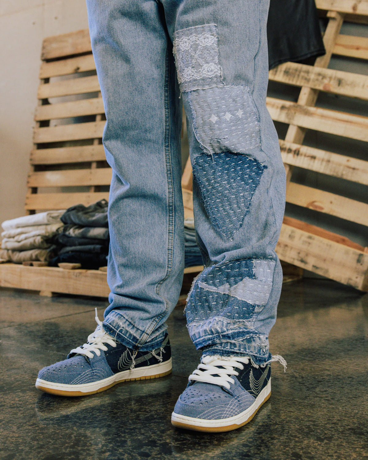 Light blue jeans with stitched denim and lace patches, worn with blue sneakers in front of wooden pallets.
