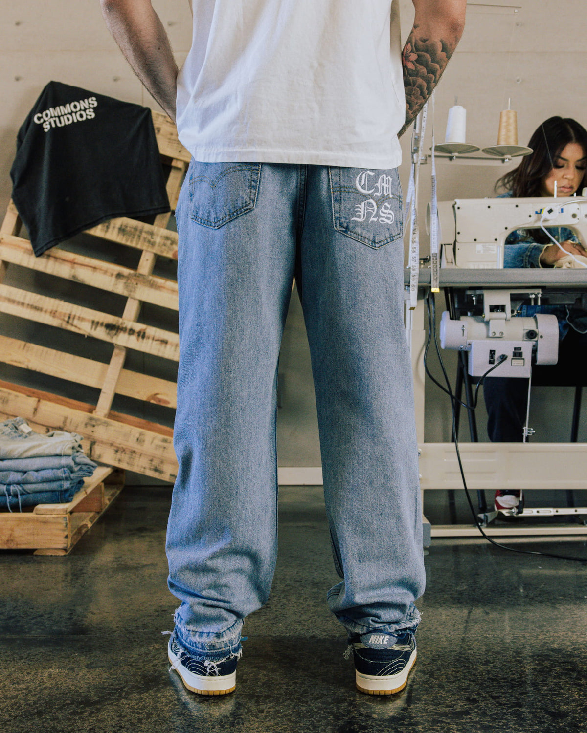 Back view of loose light blue denim jeans with white back-pocket lettering and frayed hems, worn in a sewing studio.