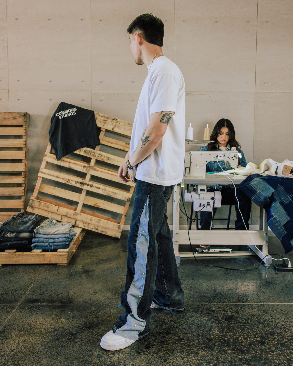Man wearing a white T-shirt and dark denim jeans with light blue studded side panels in a sewing workshop.