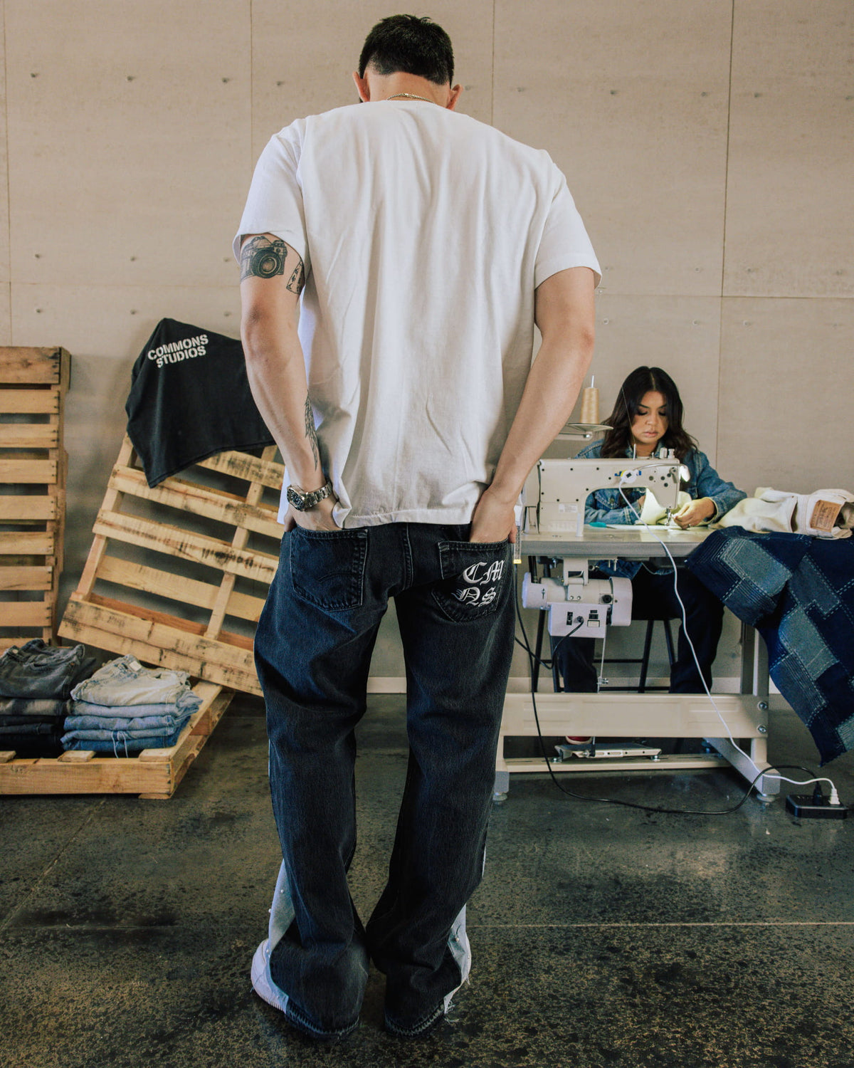 Back view of a man in a white T-shirt and dark blue wide-leg jeans with white back-pocket lettering in a sewing workshop.