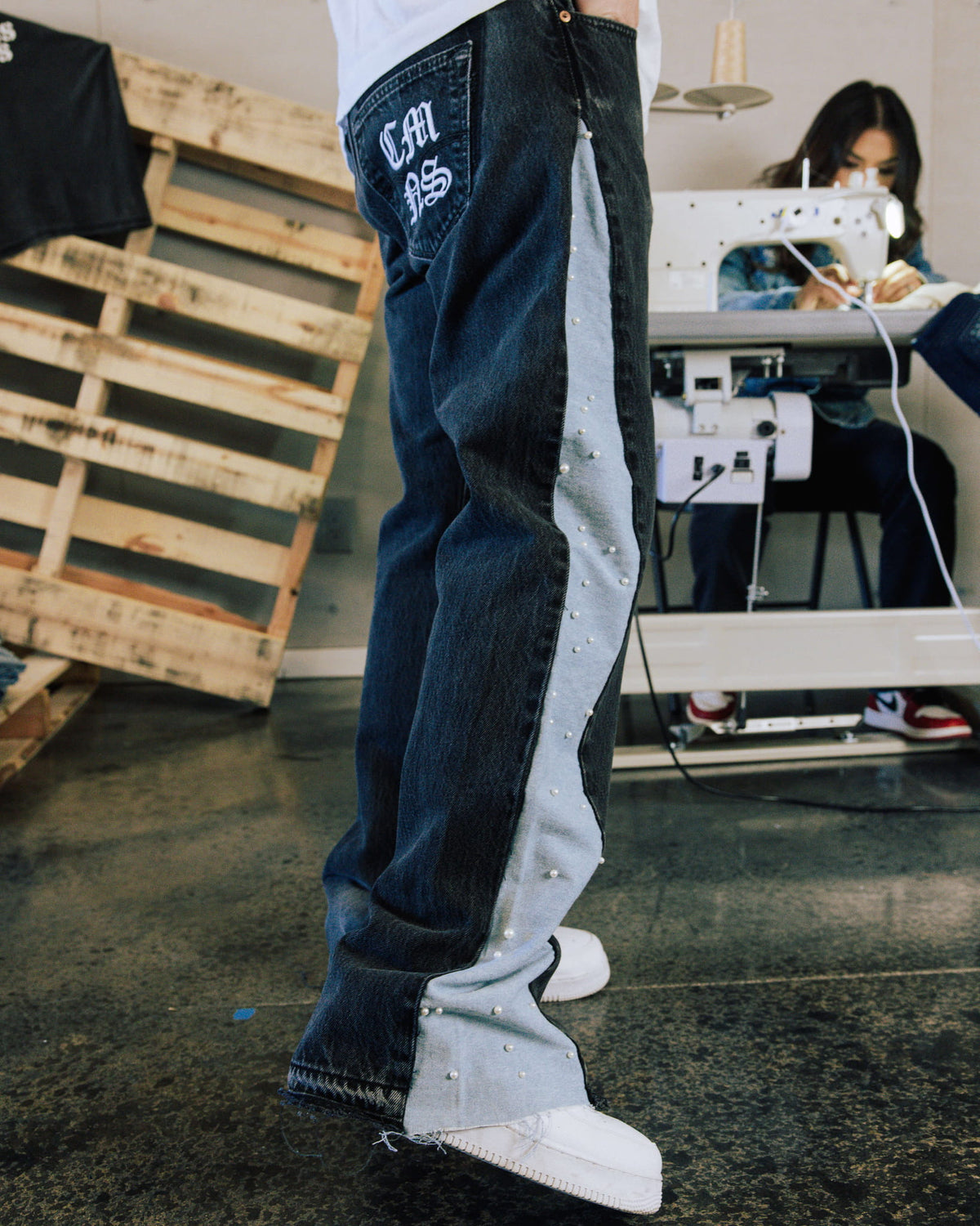 Dark denim jeans with pale blue pearl-studded side panels and white pocket lettering, worn in a sewing studio.