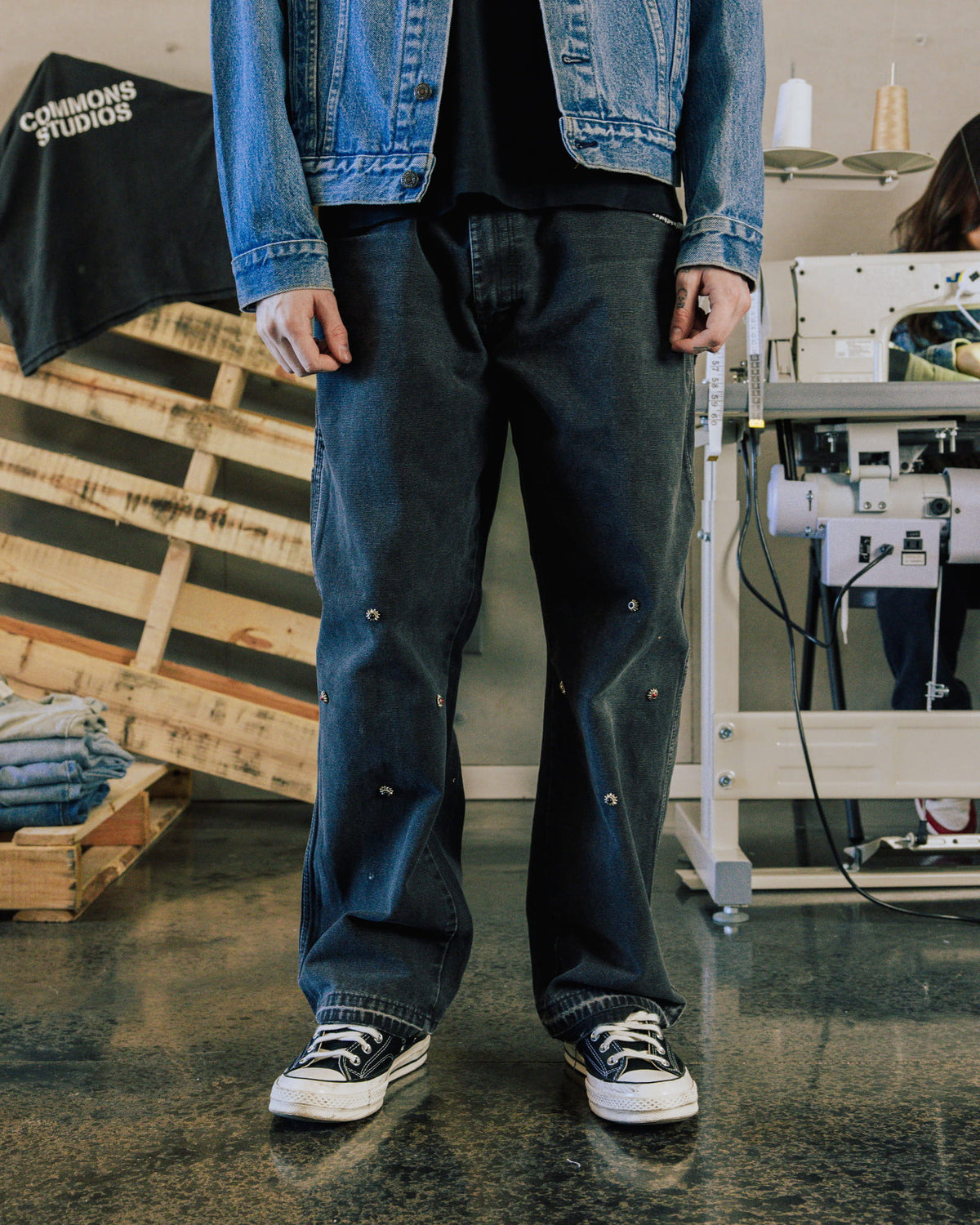 Person wearing dark wide-leg jeans with small embellishments, a blue denim jacket, and black sneakers in a sewing studio.