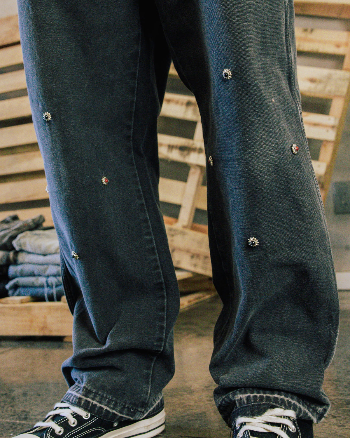 Loose blue denim jeans with small jeweled flower embellishments, worn with black canvas sneakers near wooden crates.