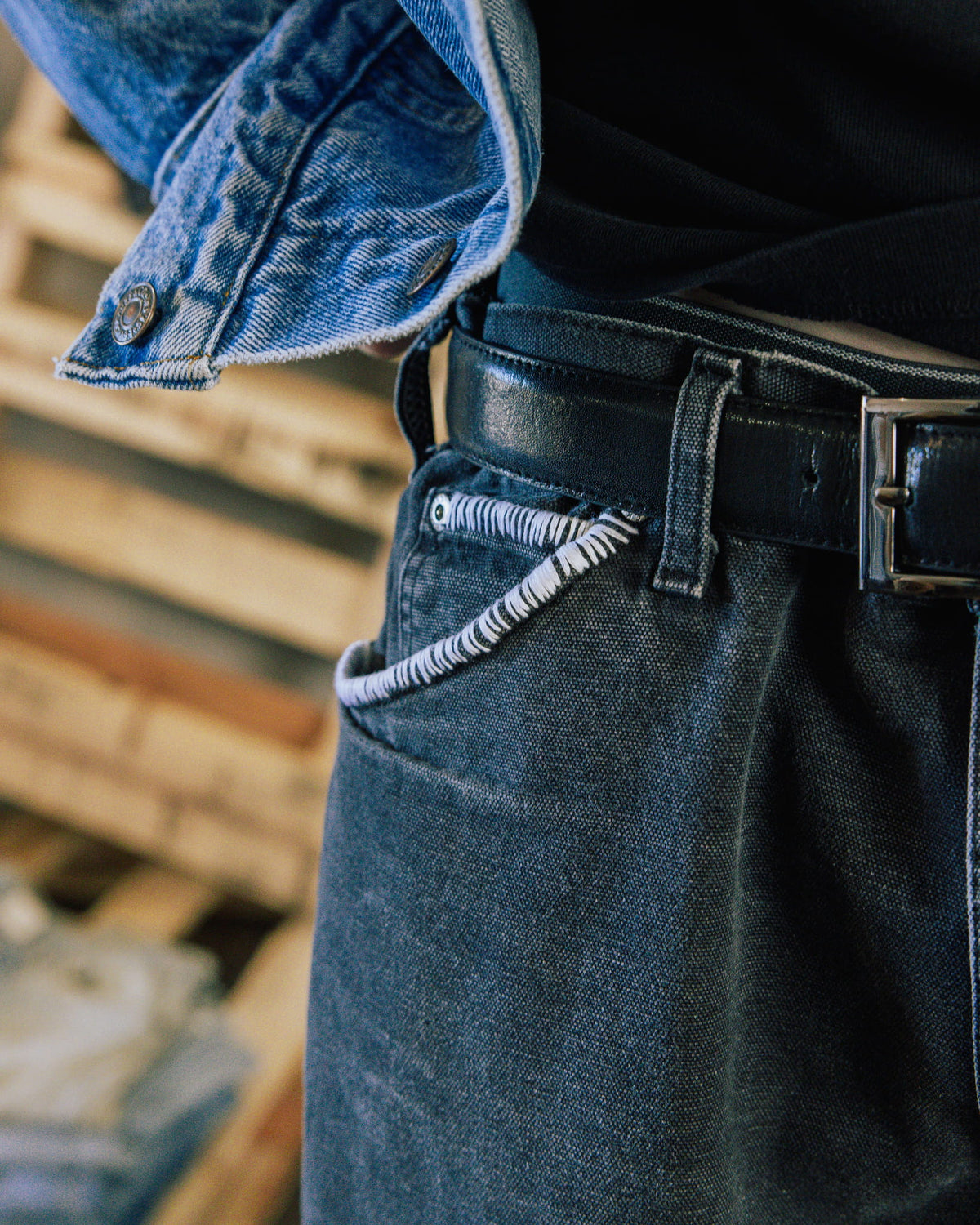 Close-up of dark jeans with white pocket stitching, a black leather belt with metal buckle, and a blue denim jacket.