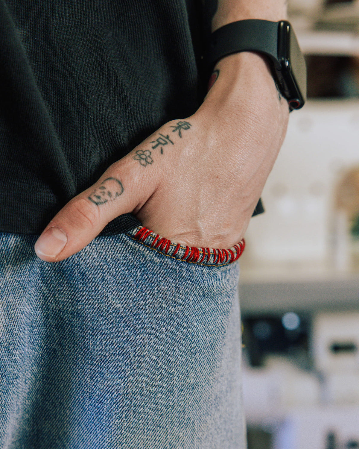 Tattooed hand tucked into a light blue denim pocket with red edge stitching, beside a black shirt and black smartwatch.