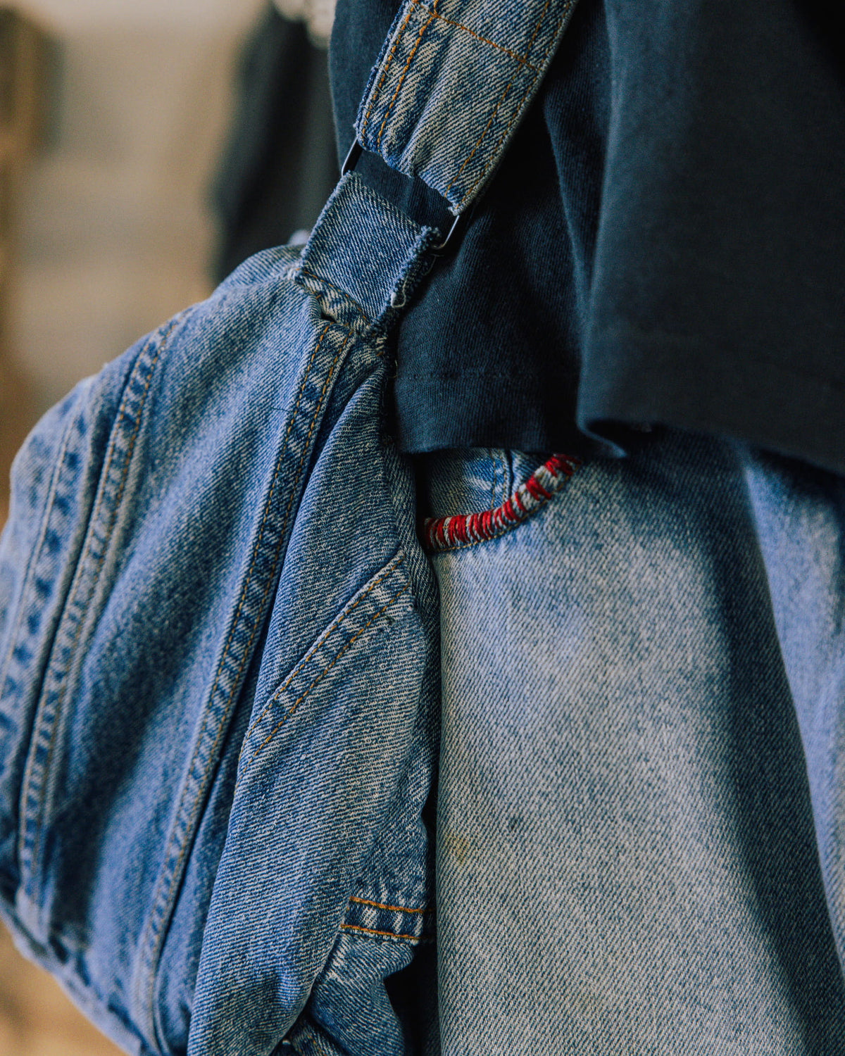 Close-up of a blue denim shoulder bag with contrast stitching and a black strap adjuster, worn over a dark shirt.