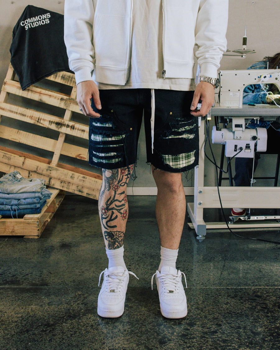 Person wearing distressed black denim shorts with plaid patches, a white zip jacket and white sneakers in a sewing studio.
