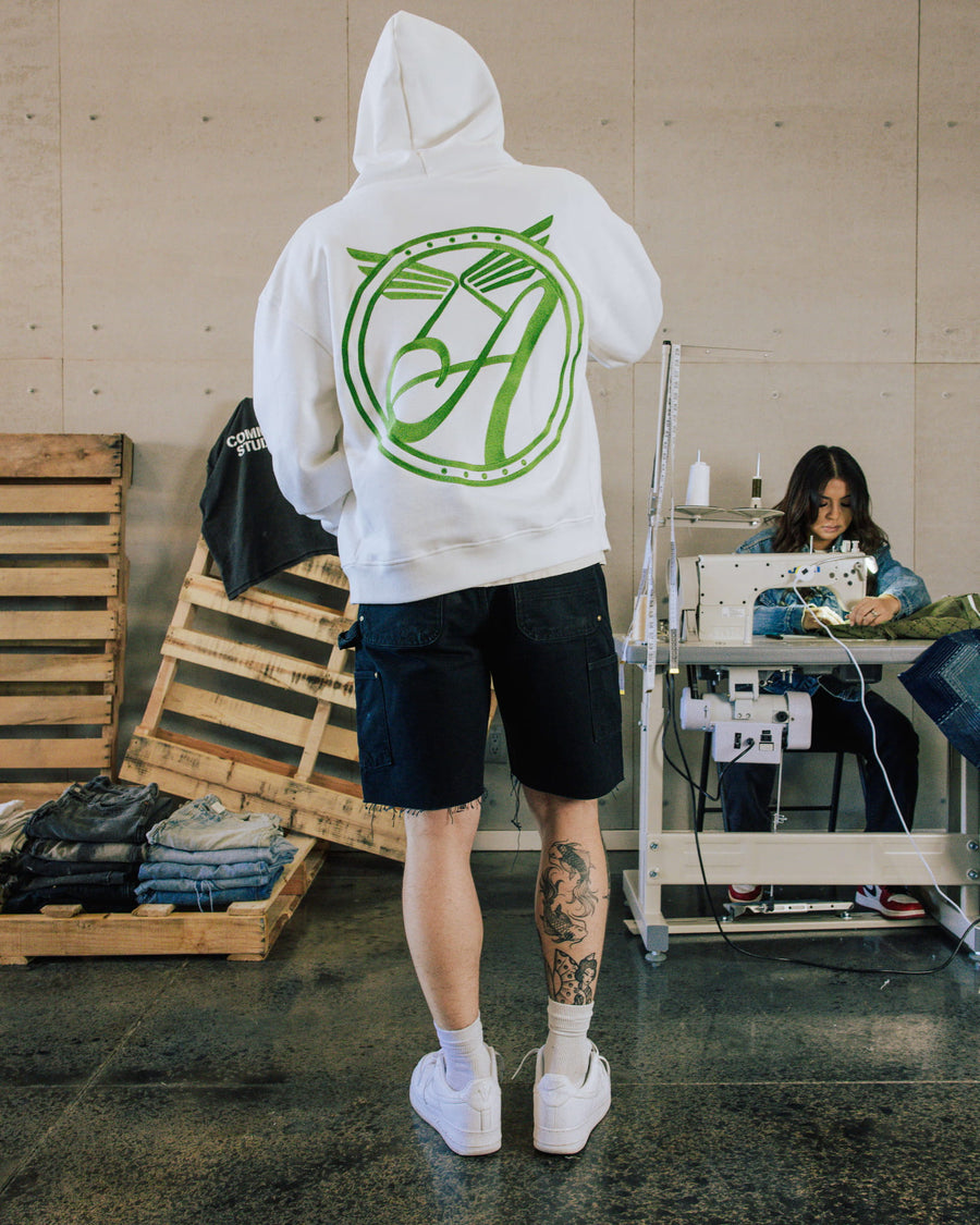 Person wearing a white hoodie with a large green back graphic and dark denim shorts in a sewing workshop.
