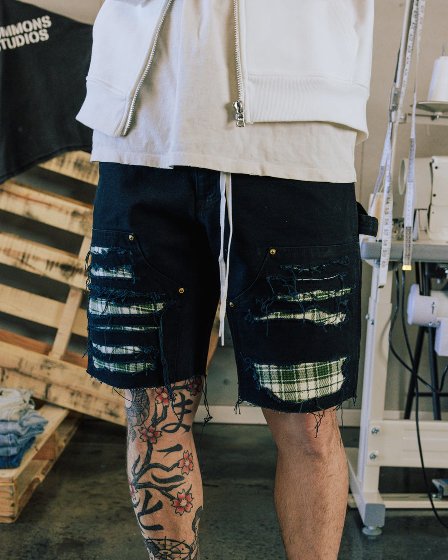 Person wearing distressed black denim shorts with green and white plaid underlays and a white drawstring in a sewing studio.