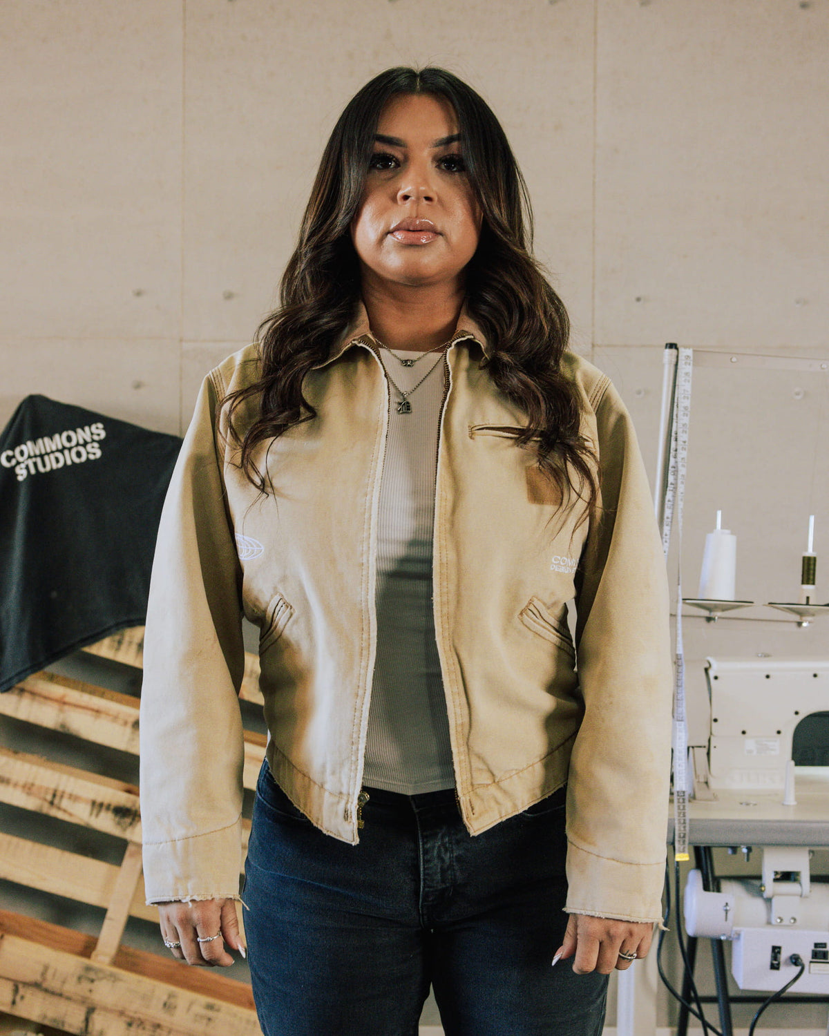 Woman wearing a tan zip-front jacket with welt pockets and raw cuffs over a white top in a sewing studio.