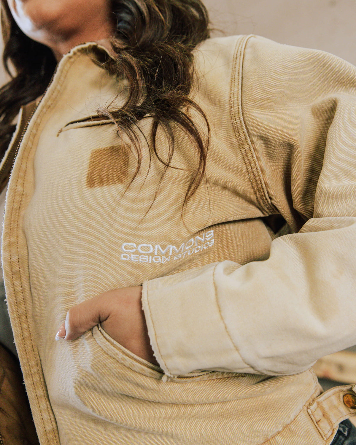 Person wearing a tan canvas zip-front jacket with white embroidered lettering, stitched seams, and a hand in the pocket.