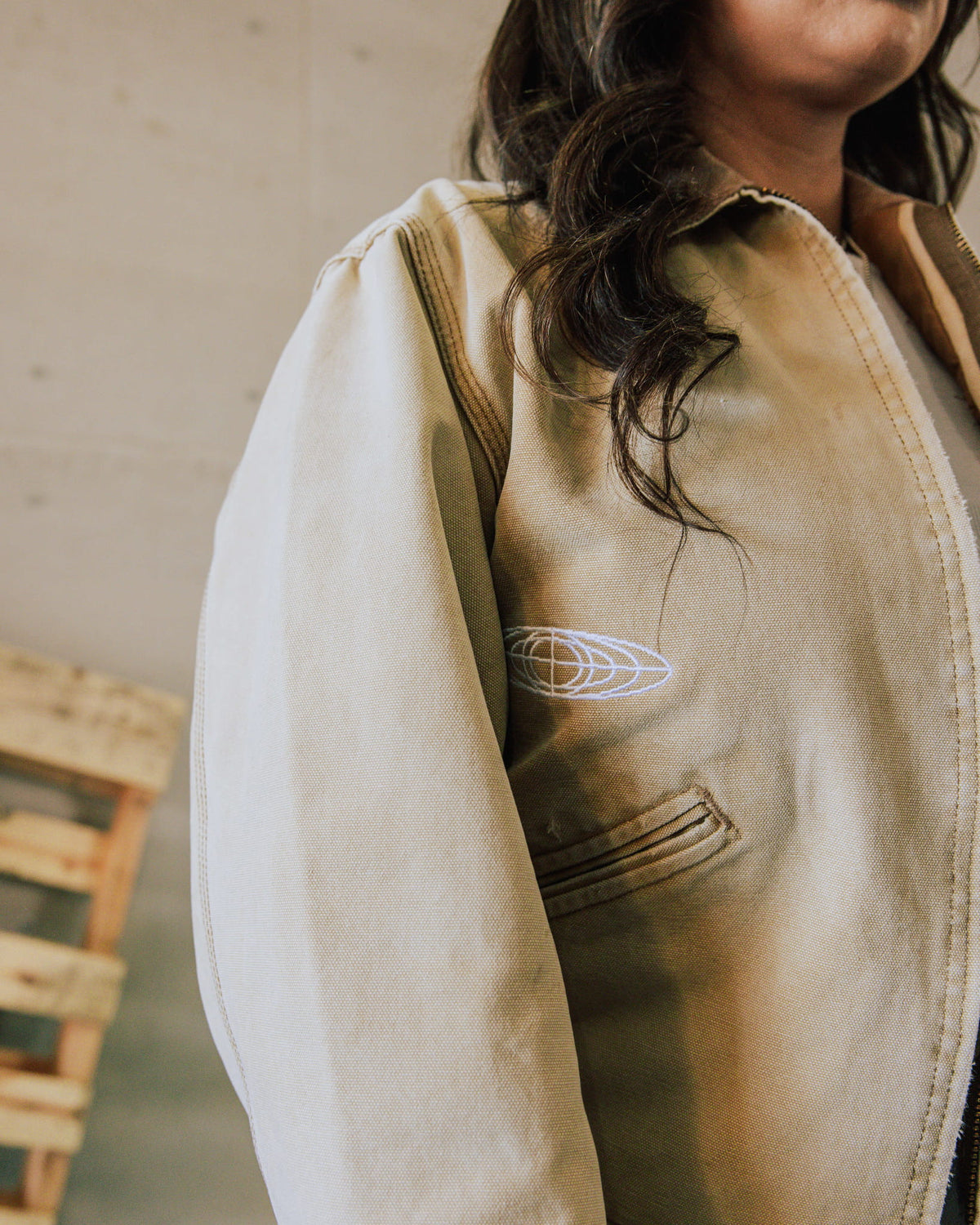 Person wearing a tan canvas zip-front jacket with contrast stitching, a welt pocket, and white globe embroidery.