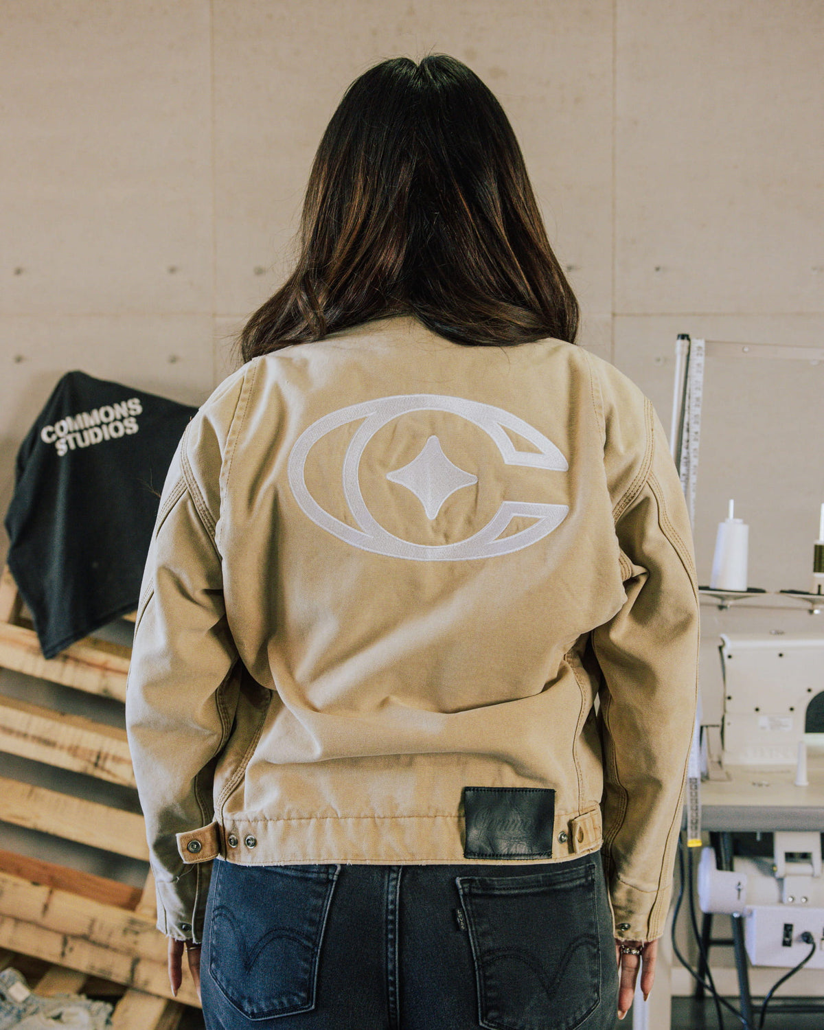 Person wearing a beige jacket with a large white embroidered back logo and dark jeans in a sewing workshop.
