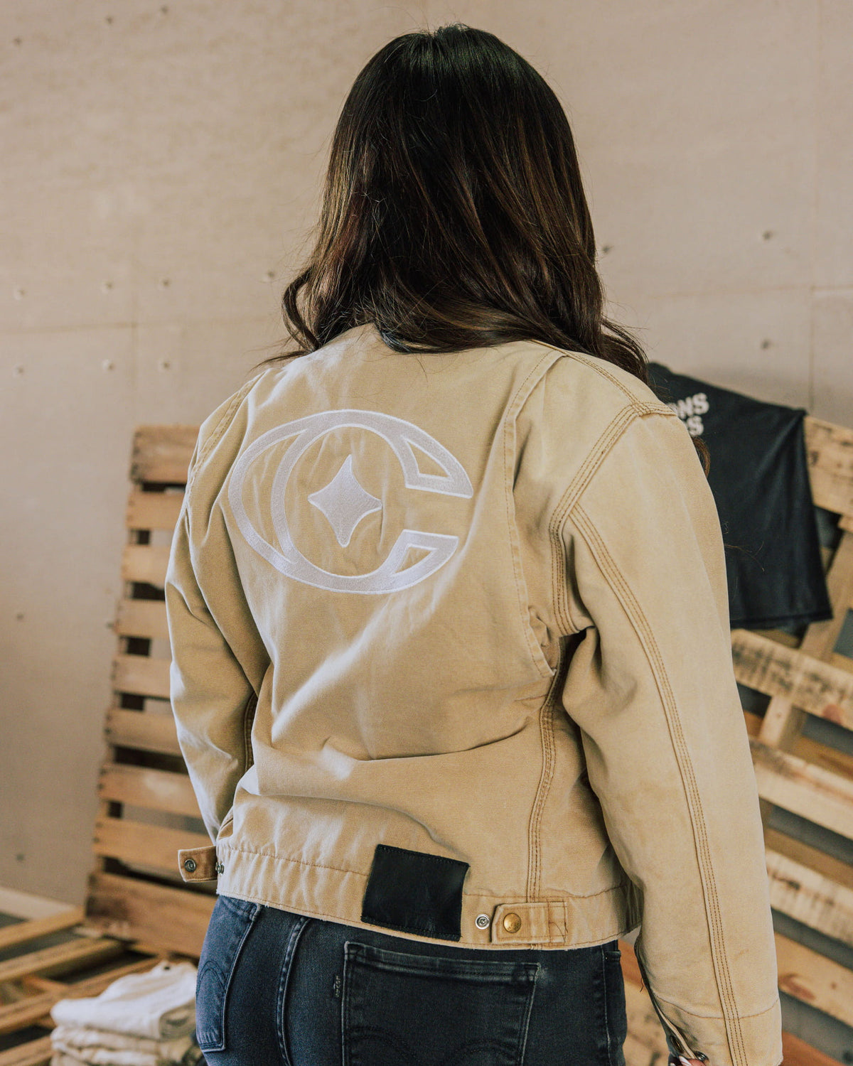 Back view of a person wearing a beige jacket with a large white embroidered logo and black hem patch beside wooden pallets.
