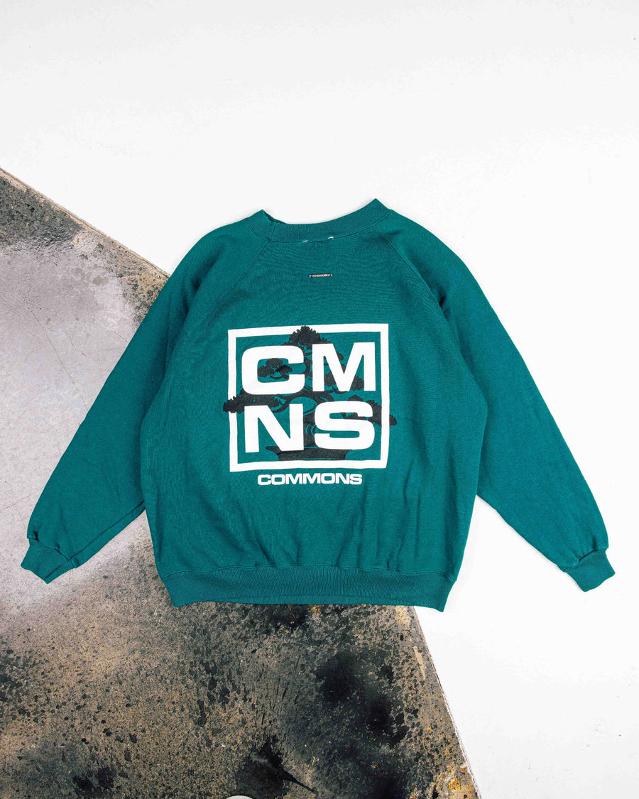 Teal crewneck sweatshirt with white CMNS lettering and a black tree graphic, laid flat on white and concrete surfaces.