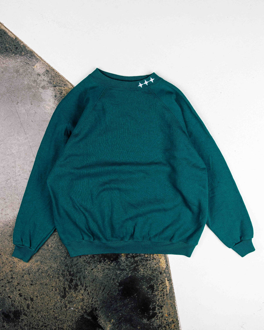 Teal crewneck sweatshirt with white star details on the collar and ribbed cuffs, laid flat on a white and concrete surface.