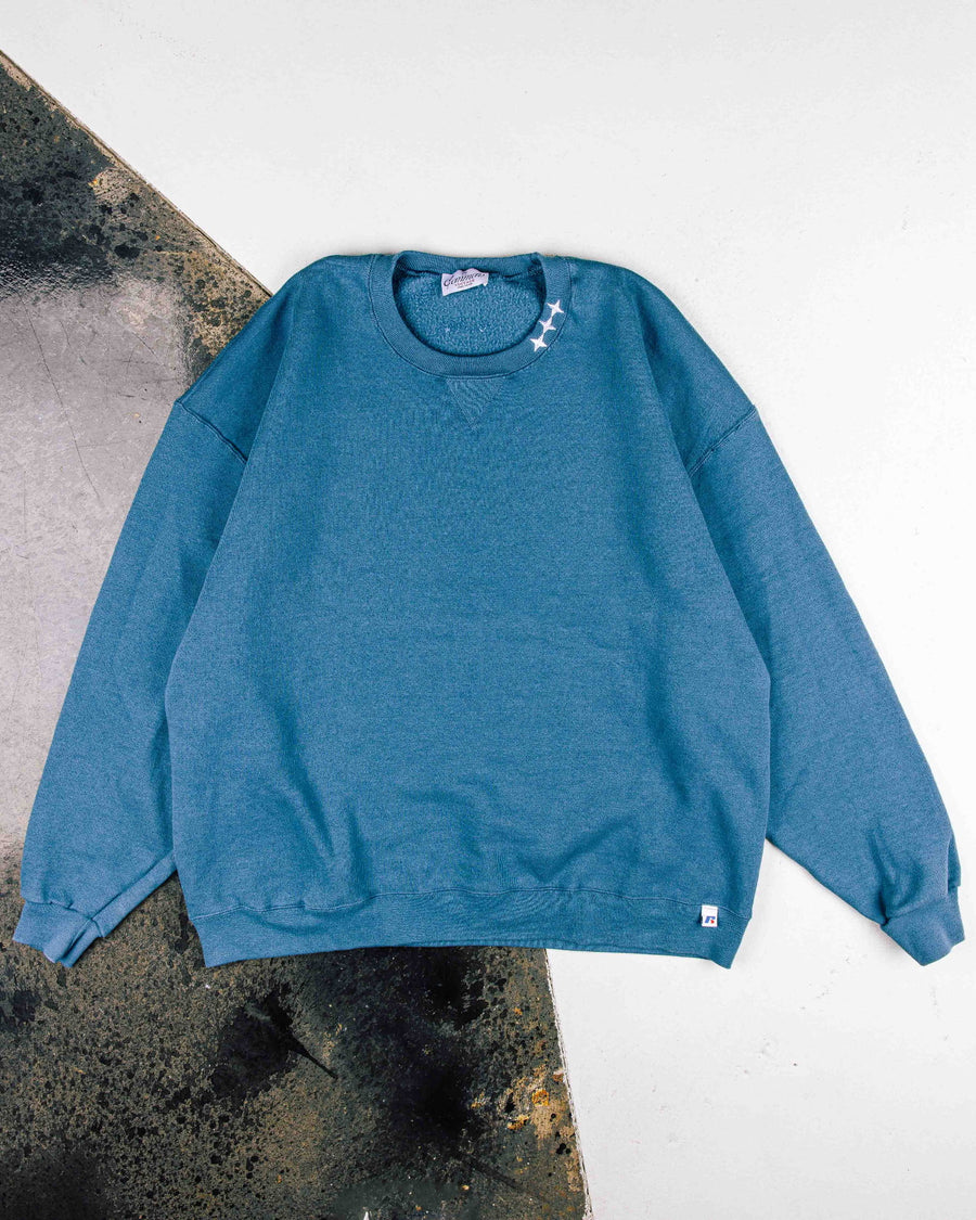 Teal crewneck sweatshirt with white star embroidery at the collar and ribbed cuffs, laid flat on a white and gray surface.