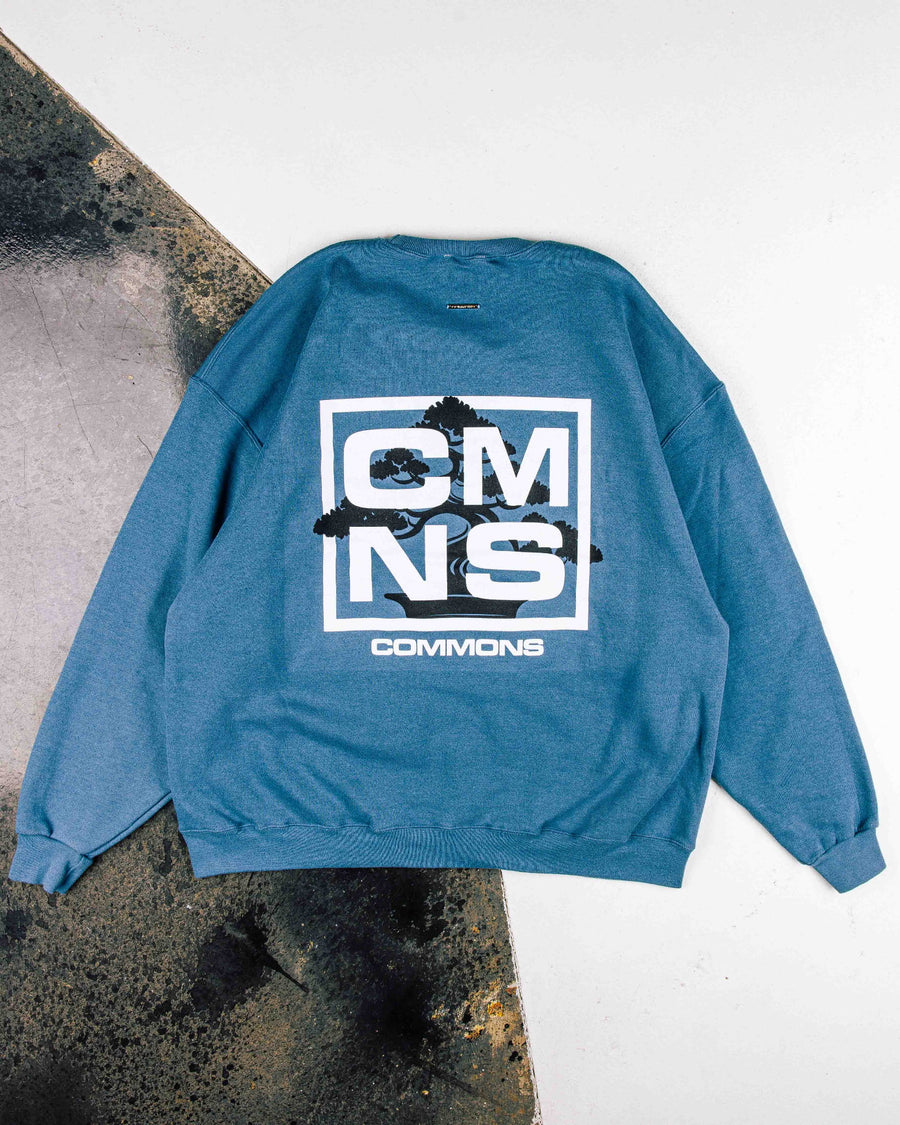 Blue crewneck sweatshirt with white CMNS lettering and black bonsai graphic on the back, laid flat on a two-tone surface.