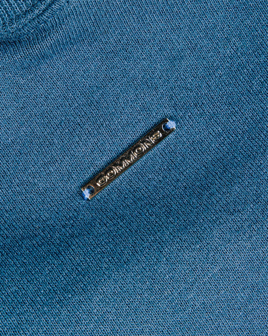 Close-up of blue knit fabric with a narrow metal COMMONS logo tag stitched at both ends.