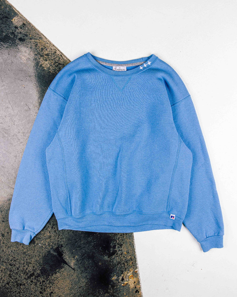 Blue crewneck sweatshirt with white stars at the collar, ribbed cuffs and hem, laid flat on a white and mottled surface.