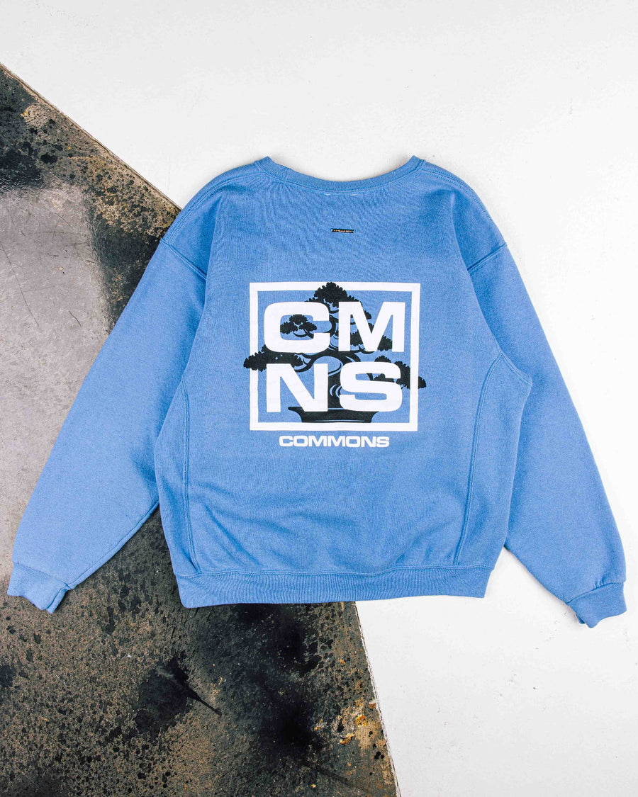 Blue crewneck sweatshirt with white CMNS lettering and black bonsai graphic, laid flat on a white and weathered surface.