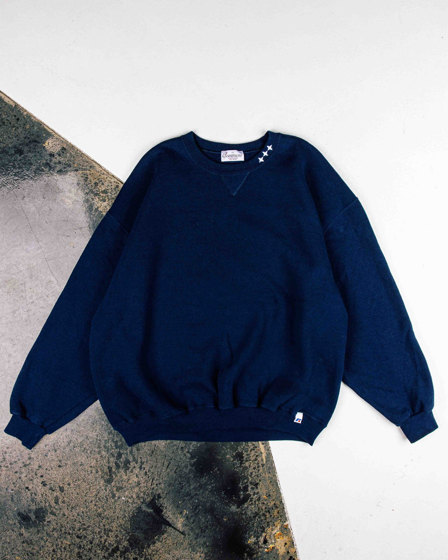 Navy crewneck sweatshirt with three white stars at the collar and ribbed cuffs, laid flat on white and worn concrete.