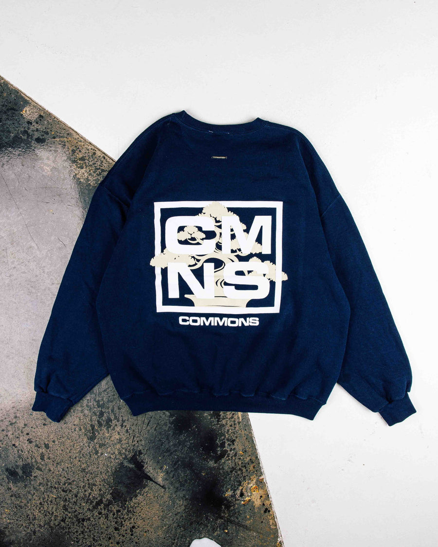 Navy sweatshirt with white CMNS lettering and a cream bonsai graphic, laid flat on a white and weathered gray surface.