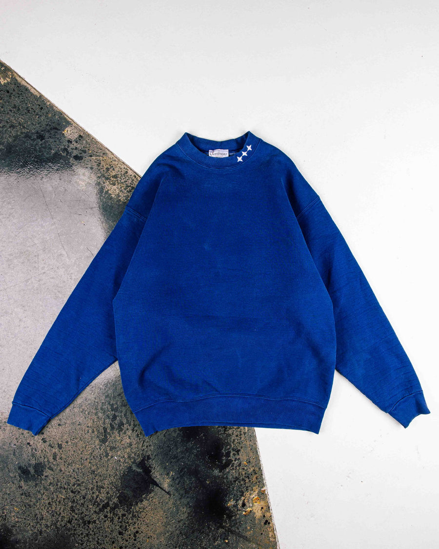 Royal blue crewneck sweatshirt with three white stars at the collar, laid flat on a white and weathered gray surface.