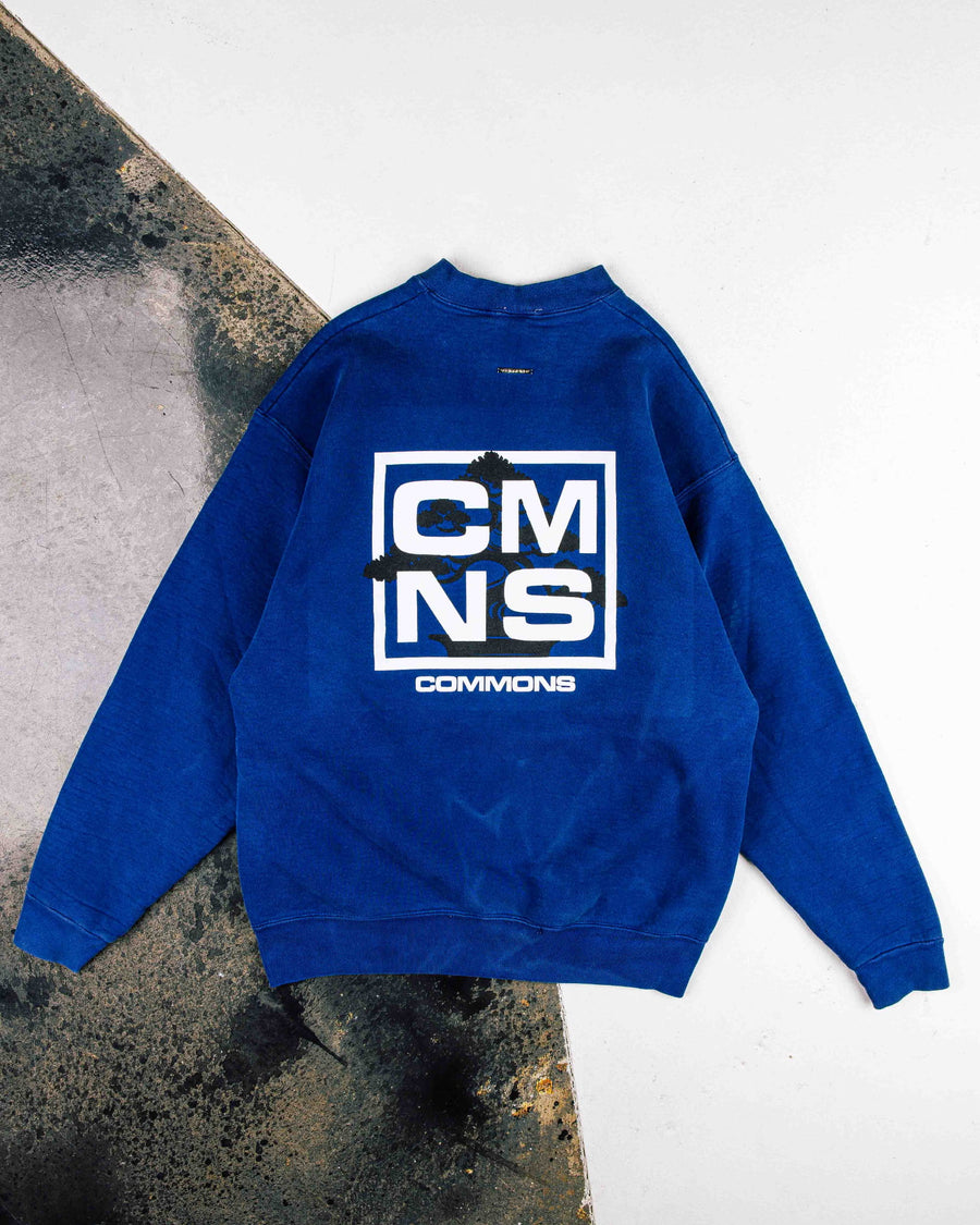 Royal blue sweatshirt with white CMNS lettering and a black tree graphic on the back, laid flat on a two-tone surface.