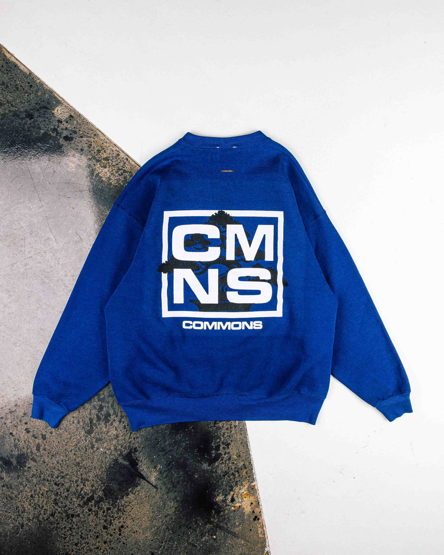 Royal blue sweatshirt with white CMNS lettering and a black tree graphic, laid flat on a white and weathered gray surface.